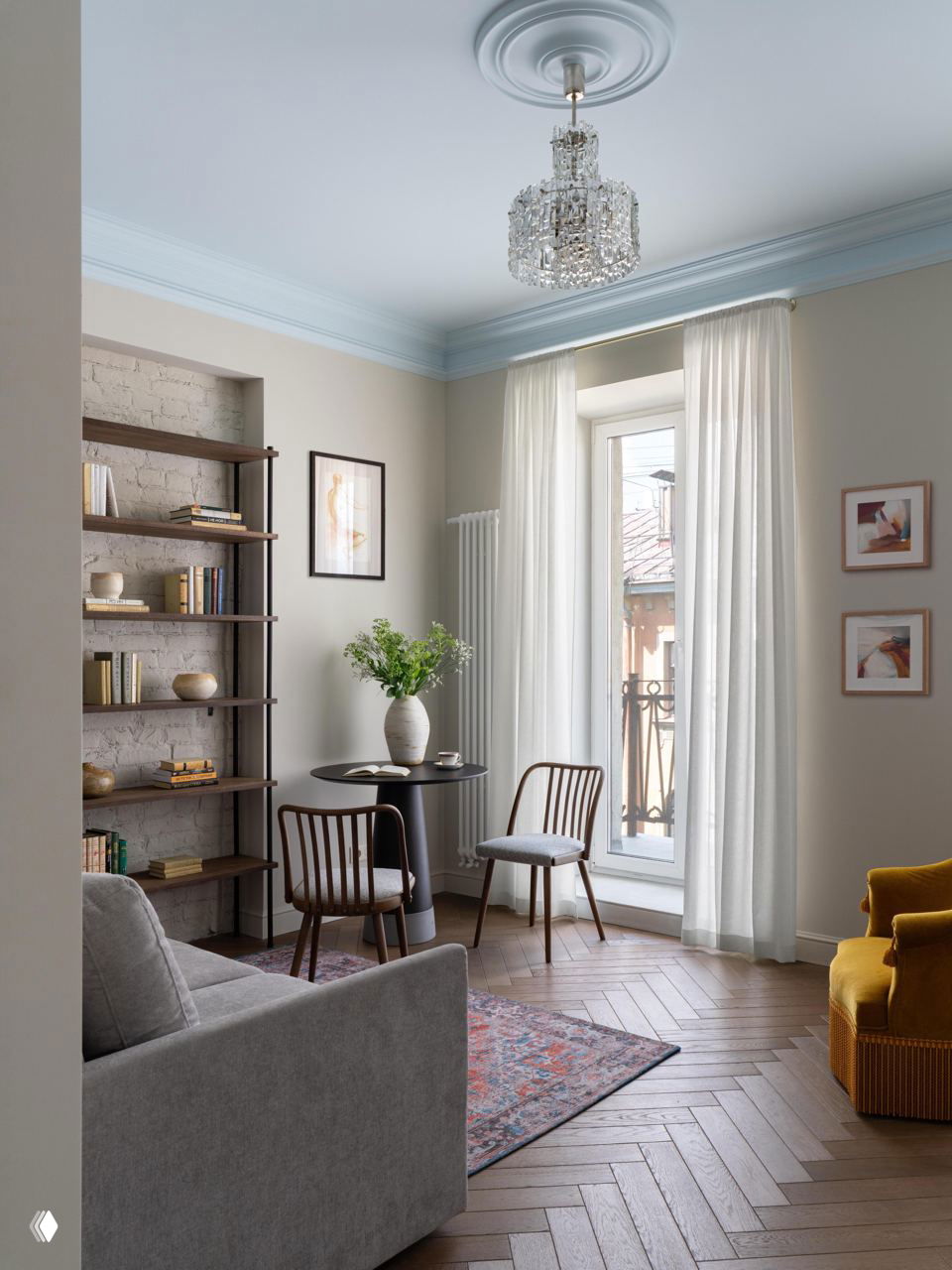 Угол гостиной с круглом столом у большого окна, тонкие шторы, светлый интерьер и хрустальная люстра на фоне неяркой пастельной палитры стен и молдингов.