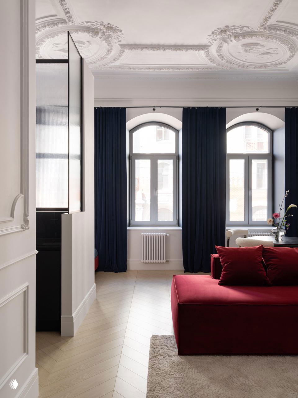 Интерьер с высокими арочными окнами, тёмными шторами и красным модульным диваном на фоне лепного потолка и светлого пола.