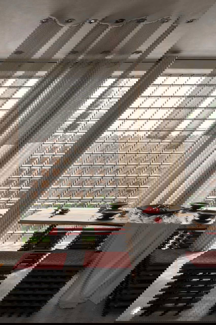 Столовая зона с длинным деревянным столом, стеклоблоковой стеной и занавесями; низкие табуреты и аккуратная композиция посуды на столе в этно-стиле.