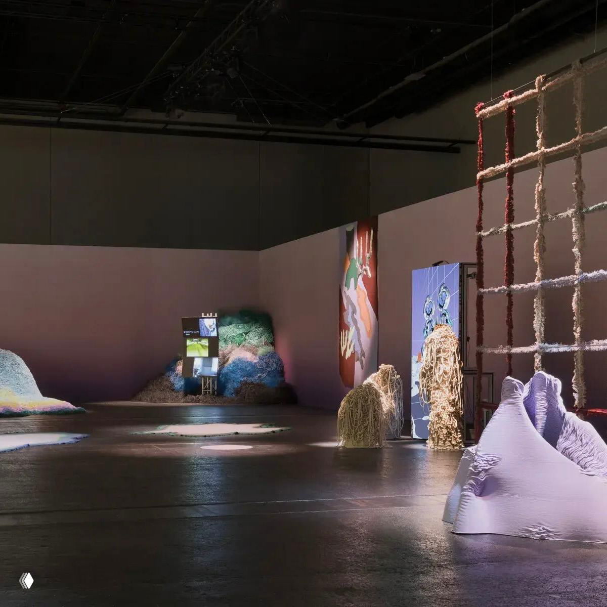 Широкий план зала Heimtextil с различными текстильными элементами: сетки, стопки переработанных нитей и мягкие формы на полу под приглушенным светом.