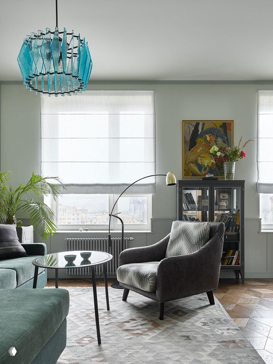 Современная гостиная с двумя окнами, бледными римскими шторами, бирюзовой стеклянной люстрой и креслом у окна в пастельной гамме.