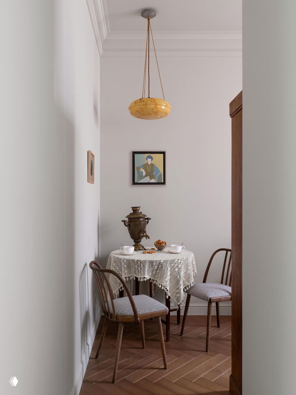 Уютный уголок с круглым столом, кружевной скатертью и самоваром, узкая проходная зона с деревянным паркетом и небольшими картинами на стенах.