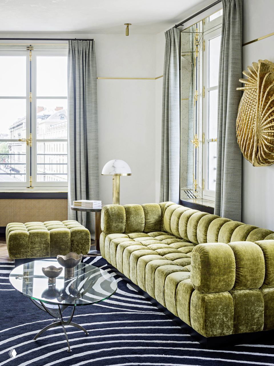 Светлая гостиная с двумя окнами, зелёным вельветовым диваном, стеклянным кофейным столом и контрастным ковром на деревянном полу.