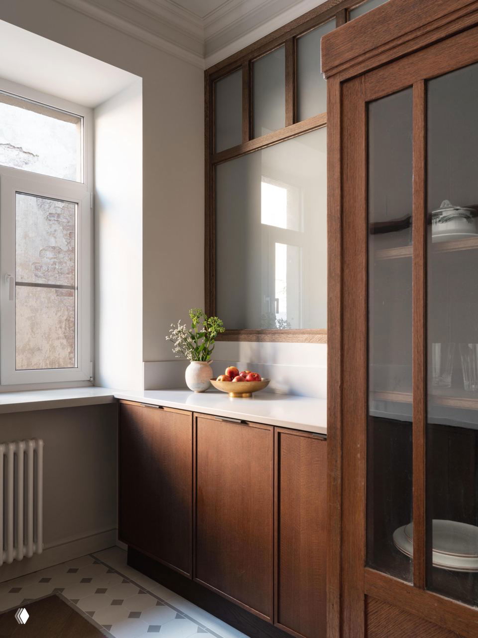 Кухонная стойка и тёмные деревянные фасады шкафа у окна, миска с фруктами на столешнице и напольная плитка у рабочей зоны в сочетании с паркетом.