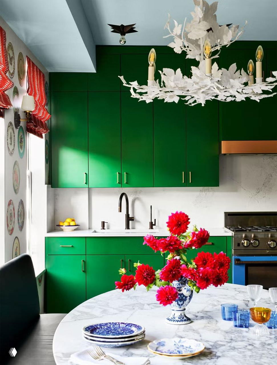 Изумрудная кухня с зелёными шкафами и мраморным столом, на котором стоит ваза с ярко‑красными цветами, посуда и стеклянные стаканы.