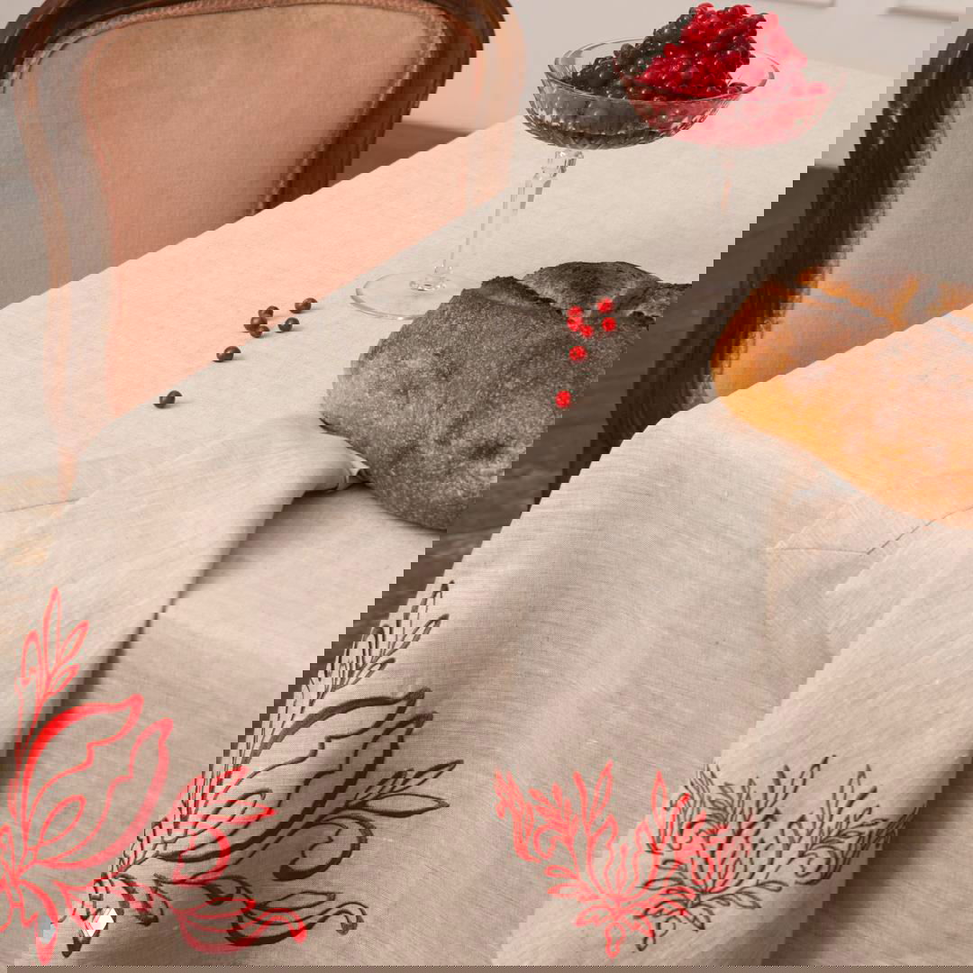 Крупный план льняной скатерти нейтрального тона с яркой красной вышивкой якобинского цветка и буханкой хлеба на углу стола.