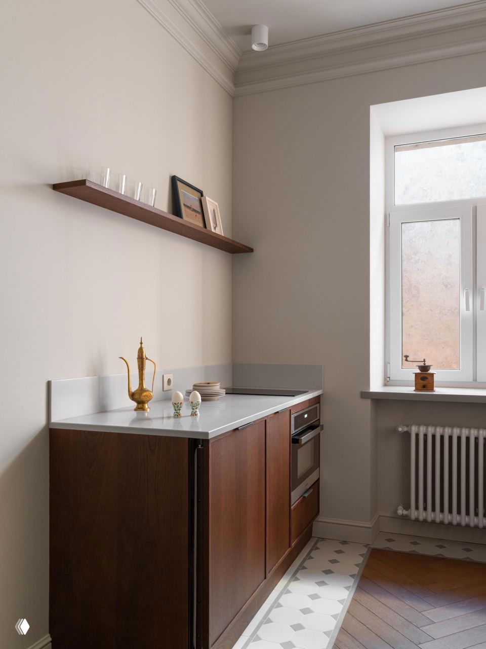 Мини-кухня с белой столешницей, встроенным духовым шкафом и тонкой деревянной полкой над рабочей зоной; свет, поступающий из высокого окна.