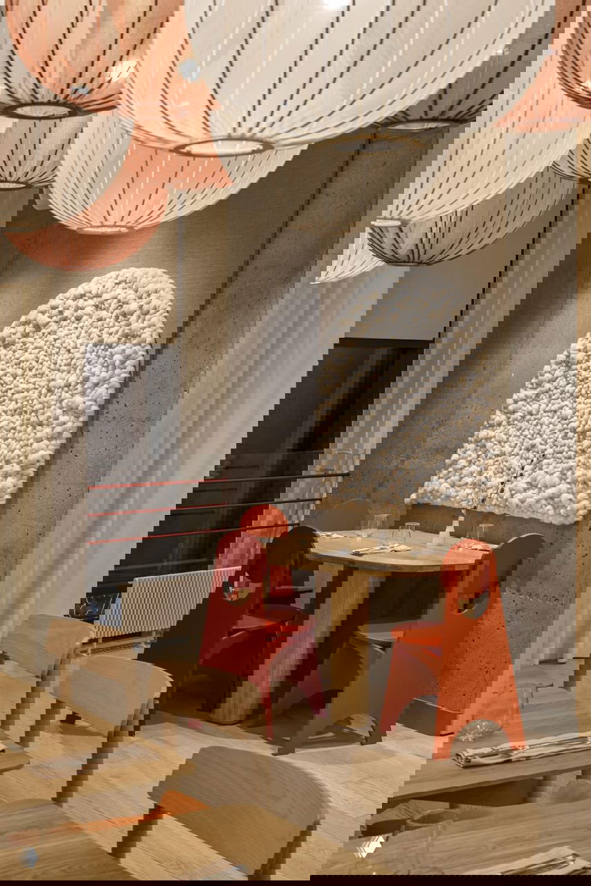 Несколько крупных тканевых подвесных абажуров над столами; на стене текстурная белая панель и яркие оранжевые стулья в контрастной точке внимания.