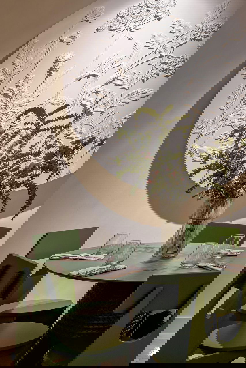 Зелёная столовая группа: круглый зелёный стол с соответствующими стульями, рельефный декоративный диск на стене и вазу с флористикой в центре.