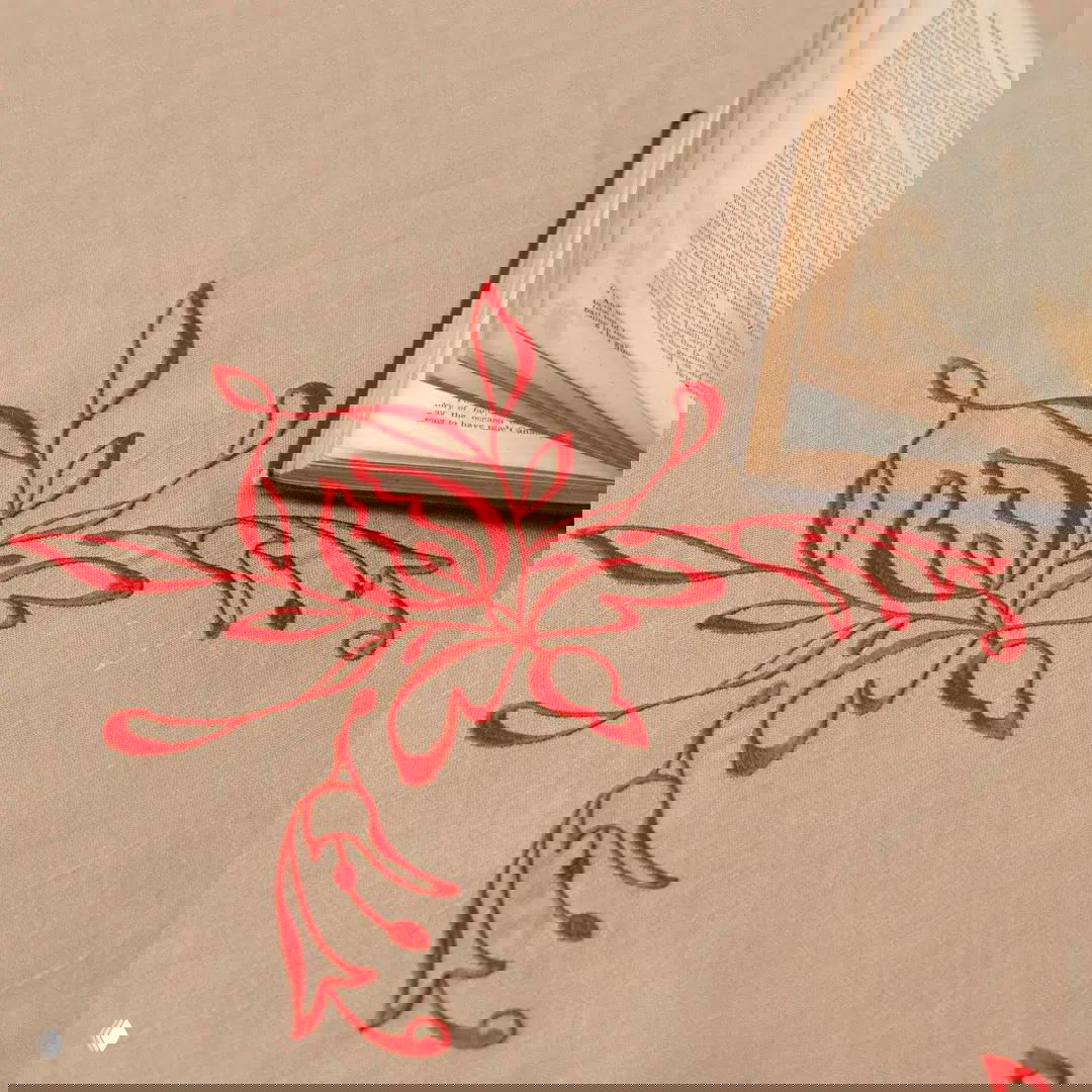 Деталь на льняной скатерти: яркая алая вышивка якобинского цветка крупным планом рядом с раскрытой книгой.