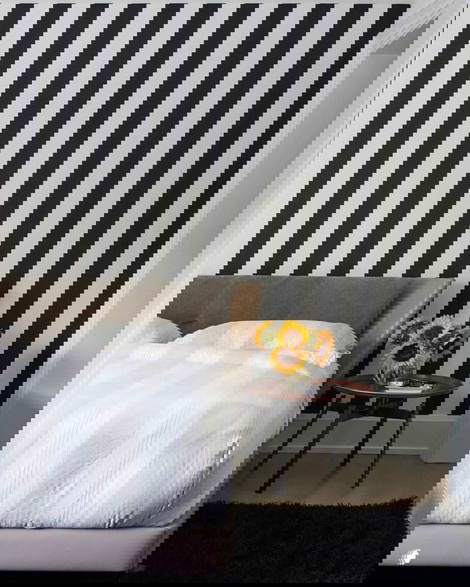Спальня с контрастными чёрно‑белыми вертикальными полосами на стене, у изголовья — светлая кровать и маленький круглый стол с букетом подсолнухов.