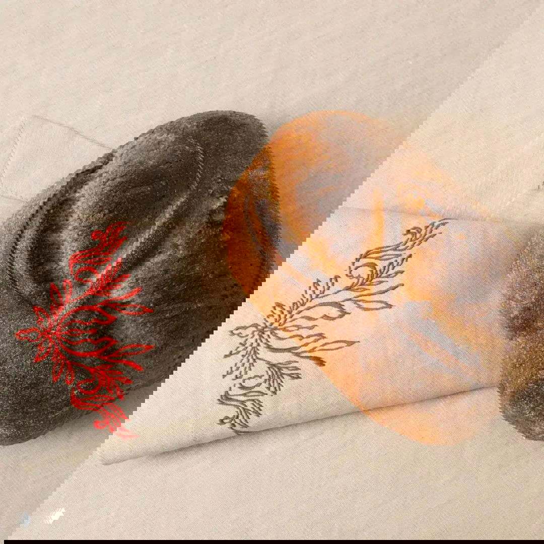 Льняной салфеточный угол с красной вышивкой и буханка хлеба, стилизованная сервировка в духе дореволюционной усадьбы.