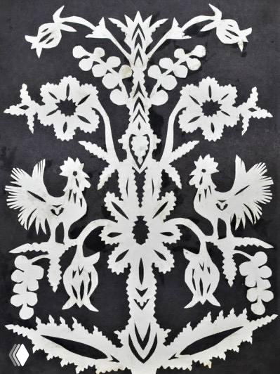 Белая вытынанка в виде цветочного мотива с двумя петухами на тёмном фоне, демонстрирует симметричный народный орнамент и ажурные вырезы.