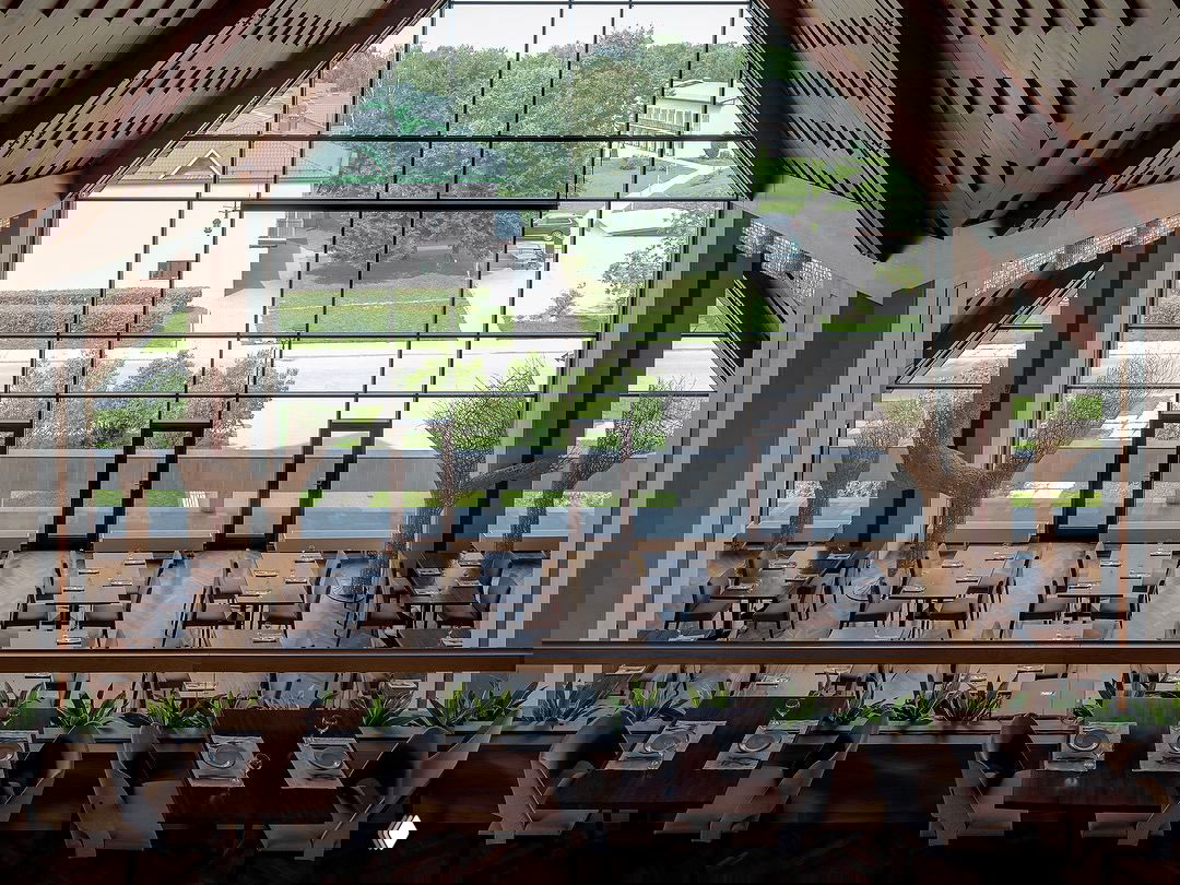 Интерьер современного кафе или ресторана с большими окнами и видом на зелёную территорию, оформленный в светлых тонах и с аккуратными посадочными местами.