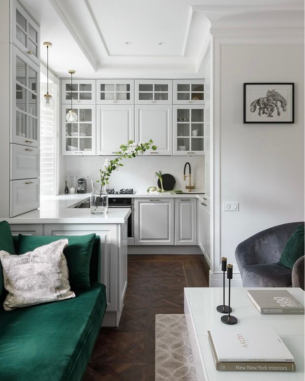 Светлая угловая кухня в классическом стиле с белыми фасадами, стеклянными вставками в верхних шкафах и зелёным диваном рядом с обеденной зоной.