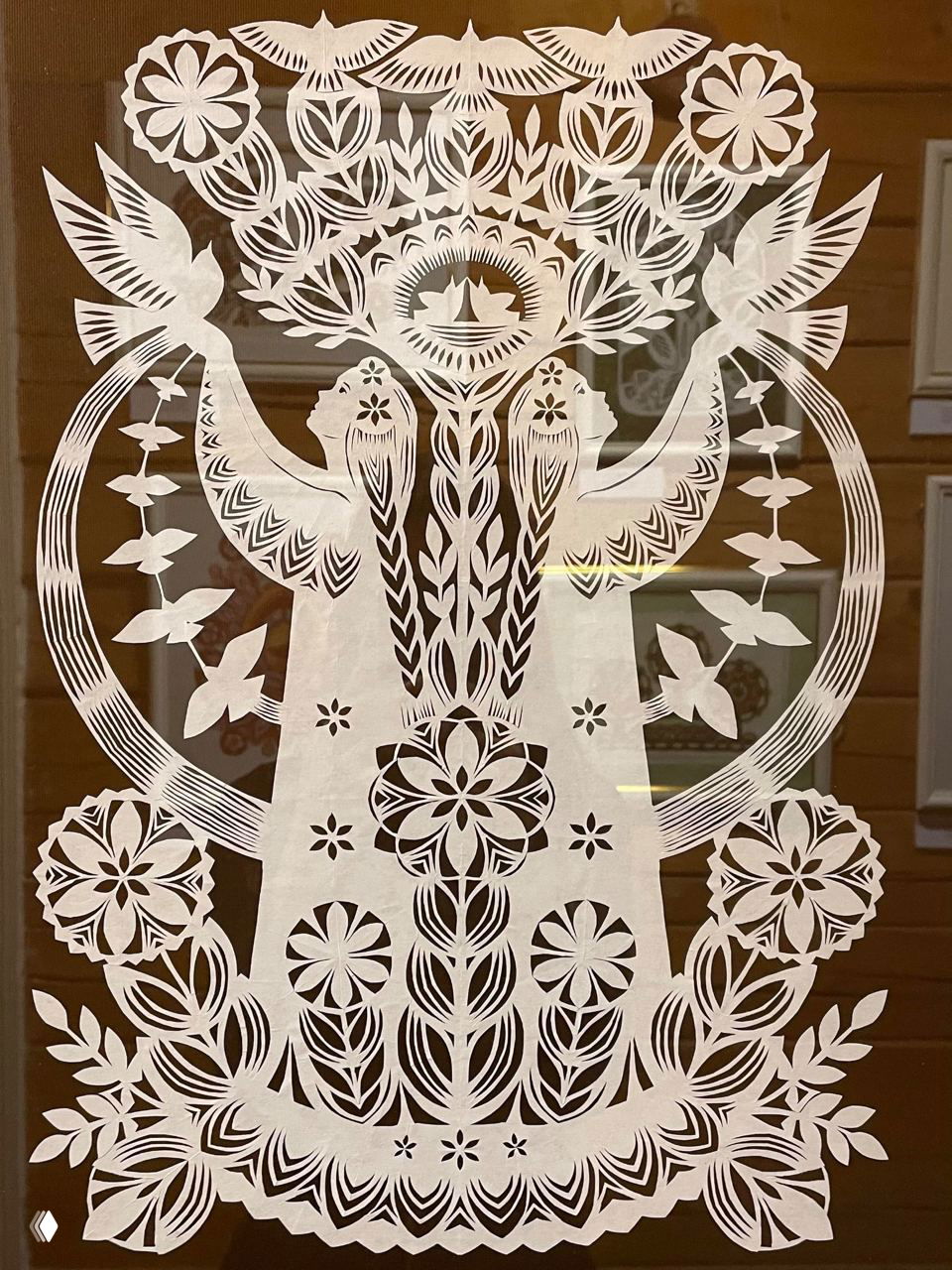 Сложная белая вытынанка на стекле с двумя фигурами в традиционных одеяниях, птицами и цветочным орнаментом, праздничное оформление окна.