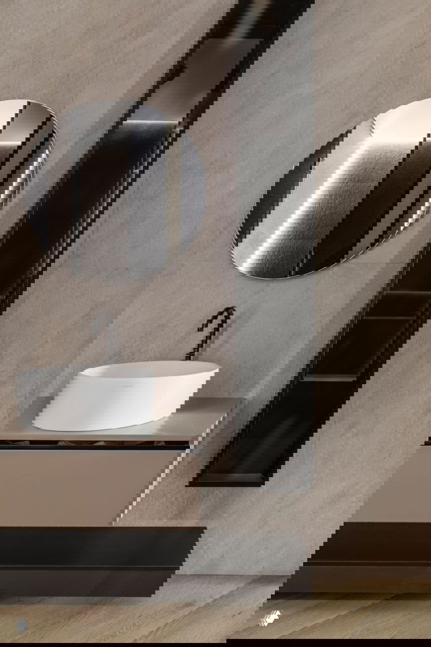 Ванная комната с круглым зеркалом над белой раковиной, бежевой плиткой и чёрными акцентами мебели и ниши в проекте Жанны Бондаренко.