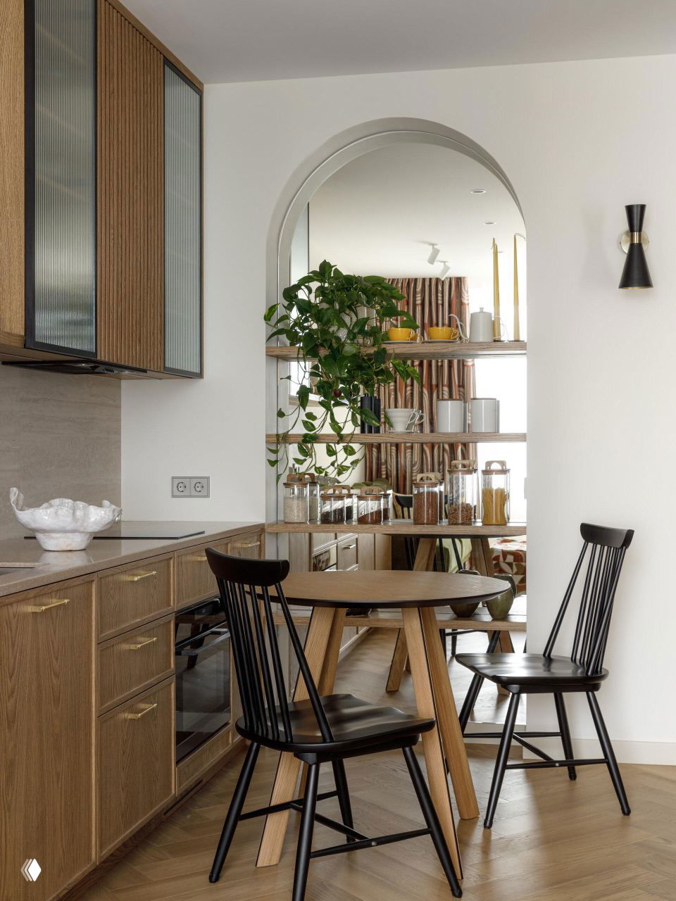Кухня-гостиная с деревянными фасадами, круглым столом и чёрными деревянными стульями; арочная ниша с полками и растением на фоне.