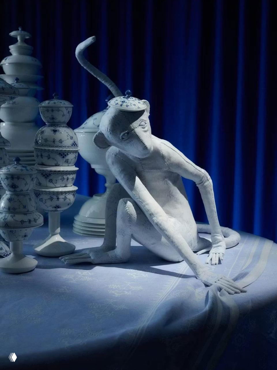 Скульптура-обезьяна из текстиля на сервированном столе рядом с фарфоровыми предметами Royal Copenhagen на синем фоне.