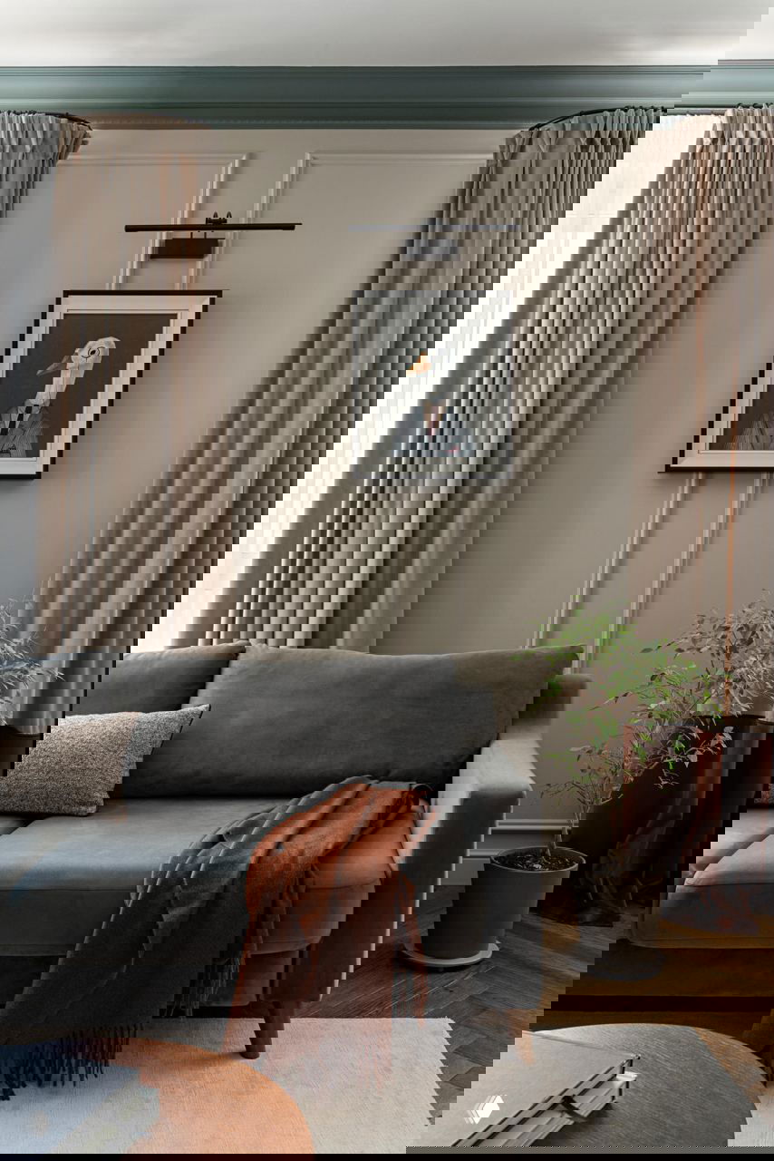 Уютная гостиная с серым диваном, бежевыми шторами, оформлением стен в молдингах и декоративной картиной с изображением птицы в раме.