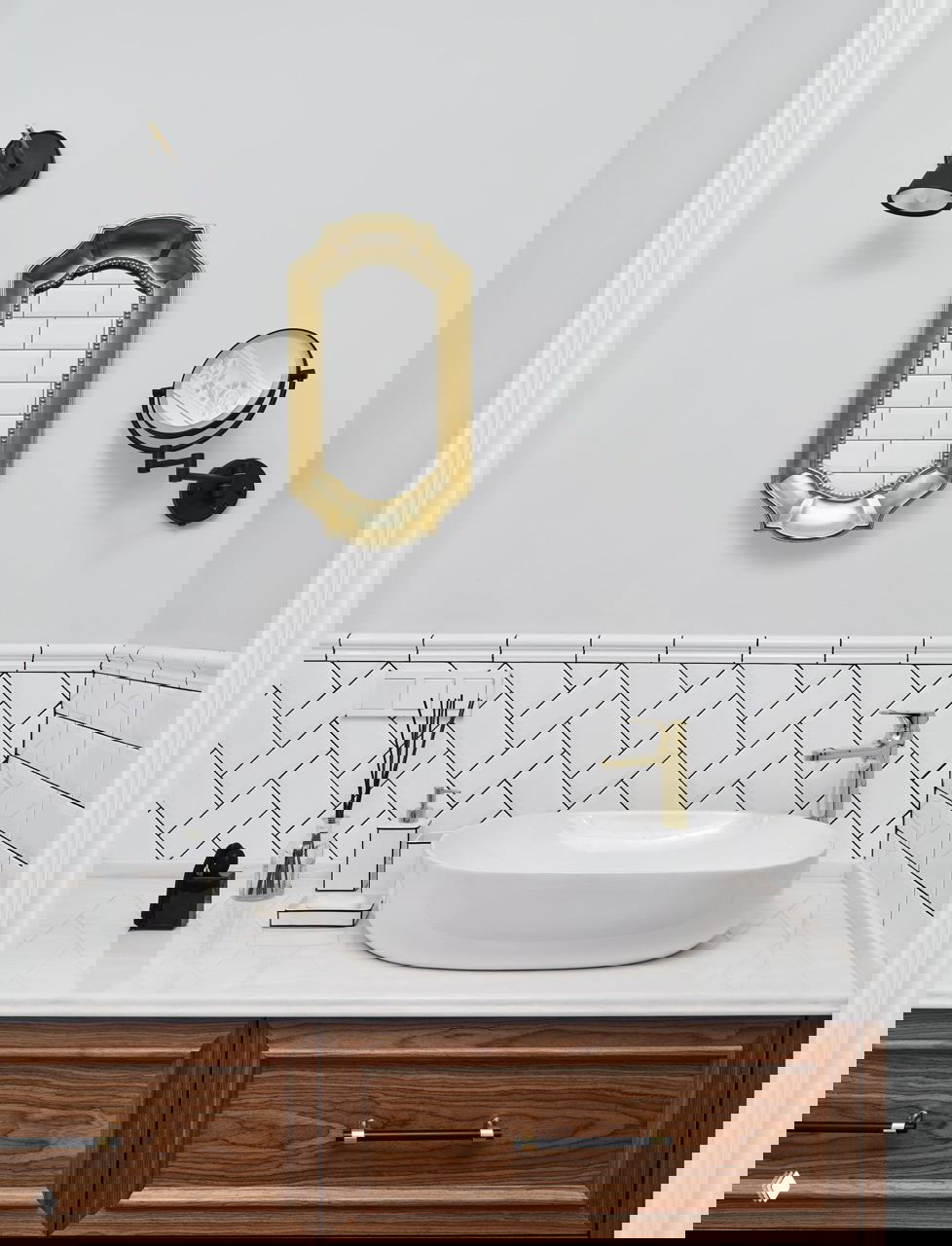 Ванная зона с белой овальной раковиной на столешнице из мрамора, декоративное золотое зеркало и плитка, выложенная ёлочкой, гармоничный ретро-акцент.