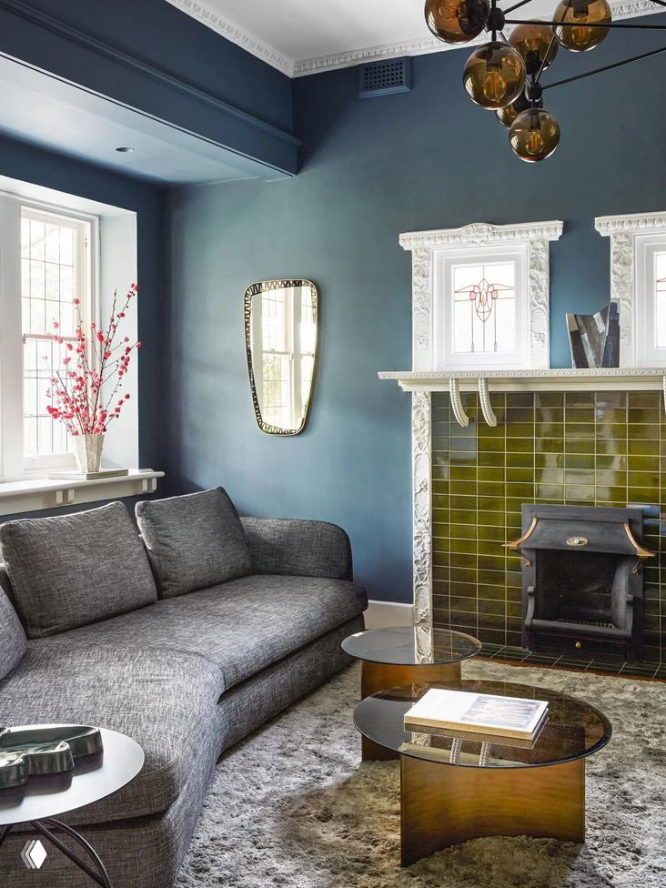 Гостиная в мельбурнском викторианском доме: глубокие насыщенно‑синие стены, лепной декоративный камин с зелёной плиткой, тёмно‑серый диван и низкие журнальные столики, создающие уют.