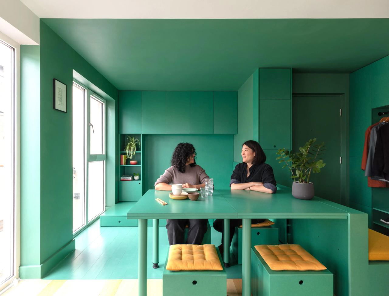 Зелёная столовая зона в мини-квартире Лондона: большой зелёный стол, встроенные шкафы и две женщины, сидящие за столом и беседующие