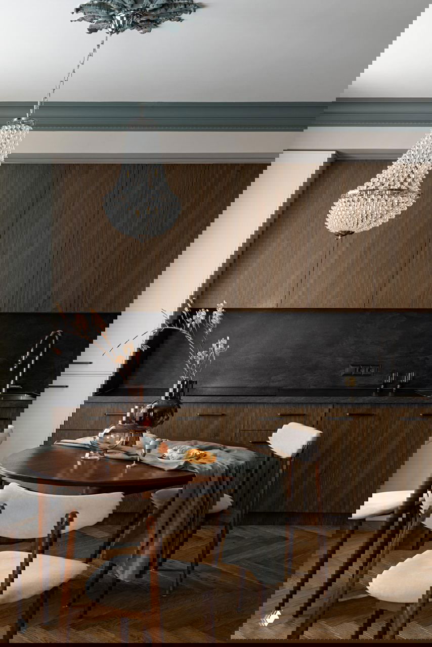 Кухонно-столовая зона: деревянные фасады шкафа, тёмная фартук-плитка, круглый стол и стулья в тёплой палитре с декоративной вазой на столе.