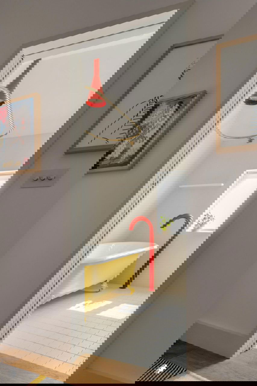 Солнечная ванная комната с жёлтой чугунной ванной и ярко-красным напольным краном, вид из дверного проёма.