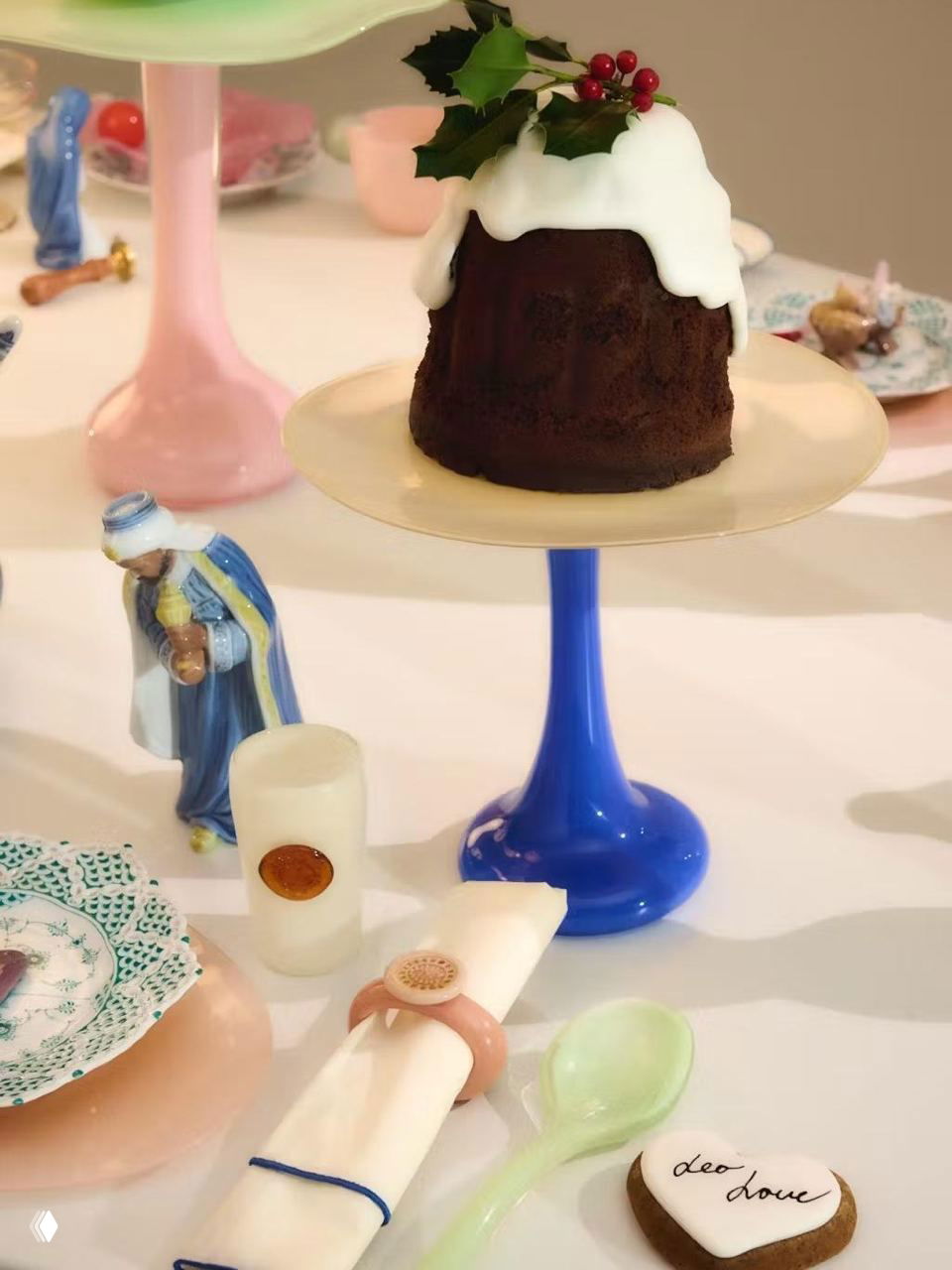 Новогодний десерт на синей подставке с белой глазурью и ягодами, рядом крошечные фигурки и столовые аксессуары на праздничном столе.