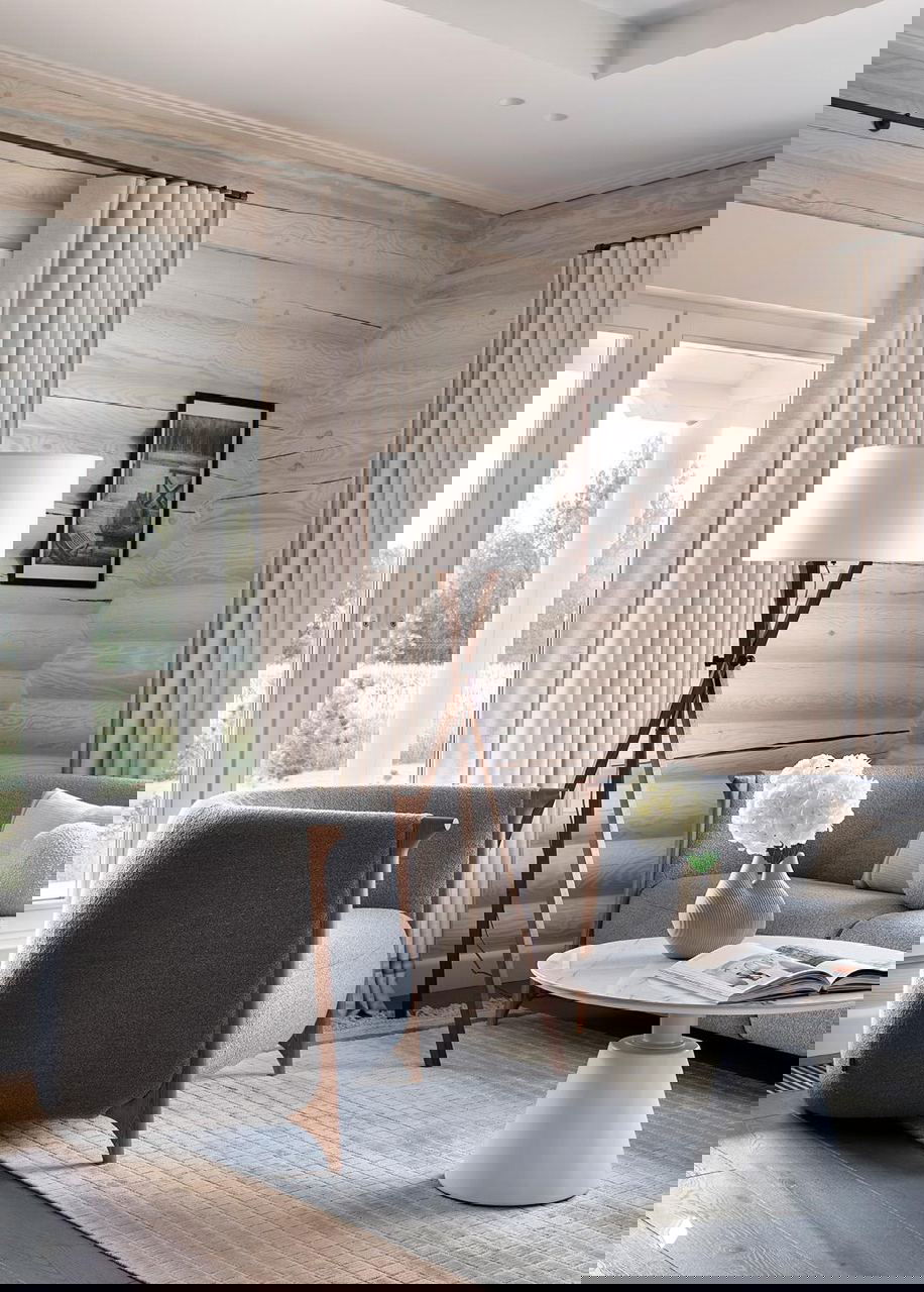 Светлая гостиная с мягкой мебелью, торшером на треноге и низким круглым столиком на фоне белёных бревенчатых стен и больших окон.