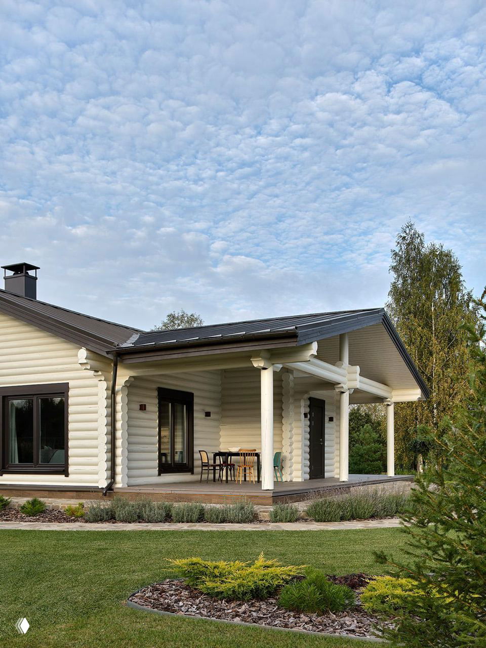 Фасад бревенчатого дома с широкой верандой и небольшой зоной отдыха на ней, вокруг ухоженный газон и декоративные посадки в Подмосковье.