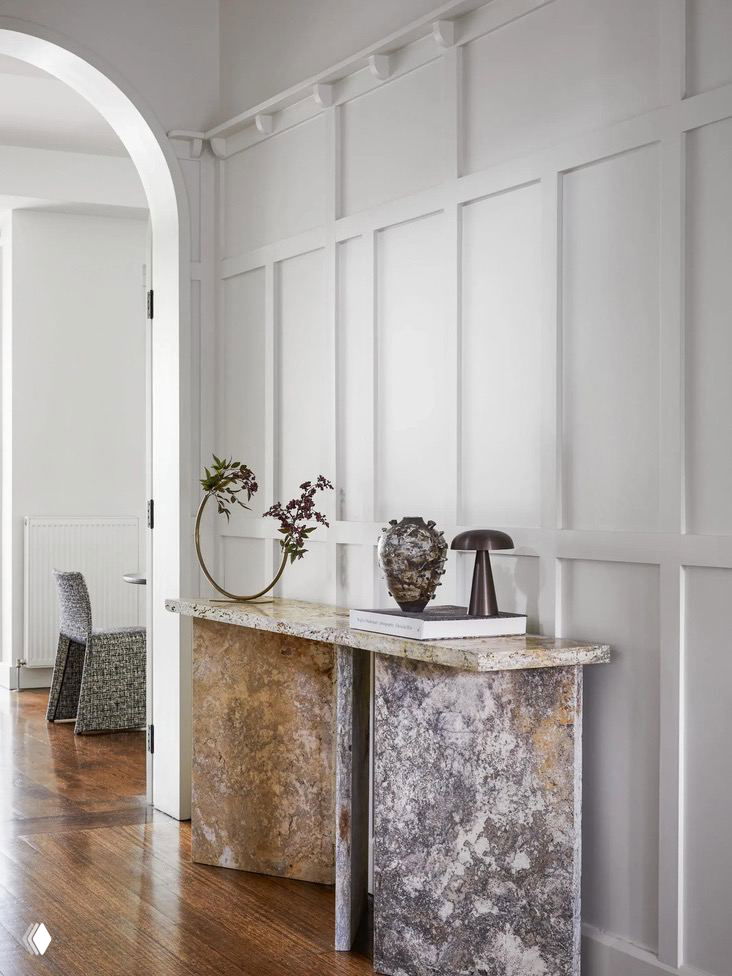Коридор и консоль в доме Малверн: молдинги и декоративные панели на стенах, паркетный пол и скульптурная консоль из натурального камня на фоне приглушённой светлой палитры.