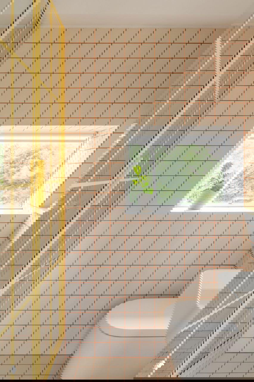 Туалетная комната с плиткой в мелкую сетку и жёлтой рамой душевой кабины, окно с видом на зелёную зелень снаружи.