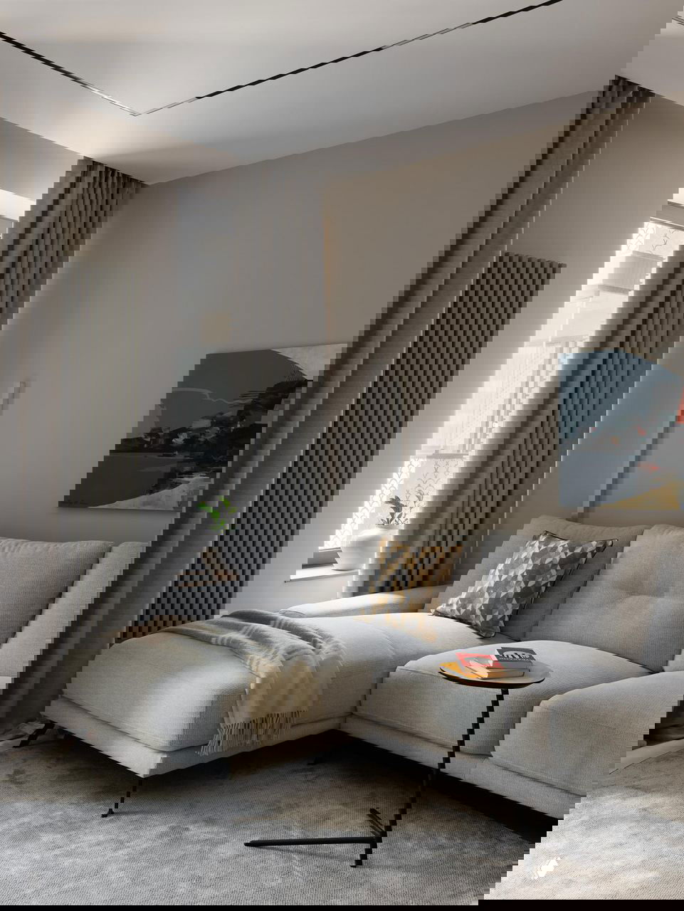 Уютная зона отдыха с угловым светлым диваном, декоративной подушкой, пледом и низким столиком на мягком ковре в нейтральных тонах.