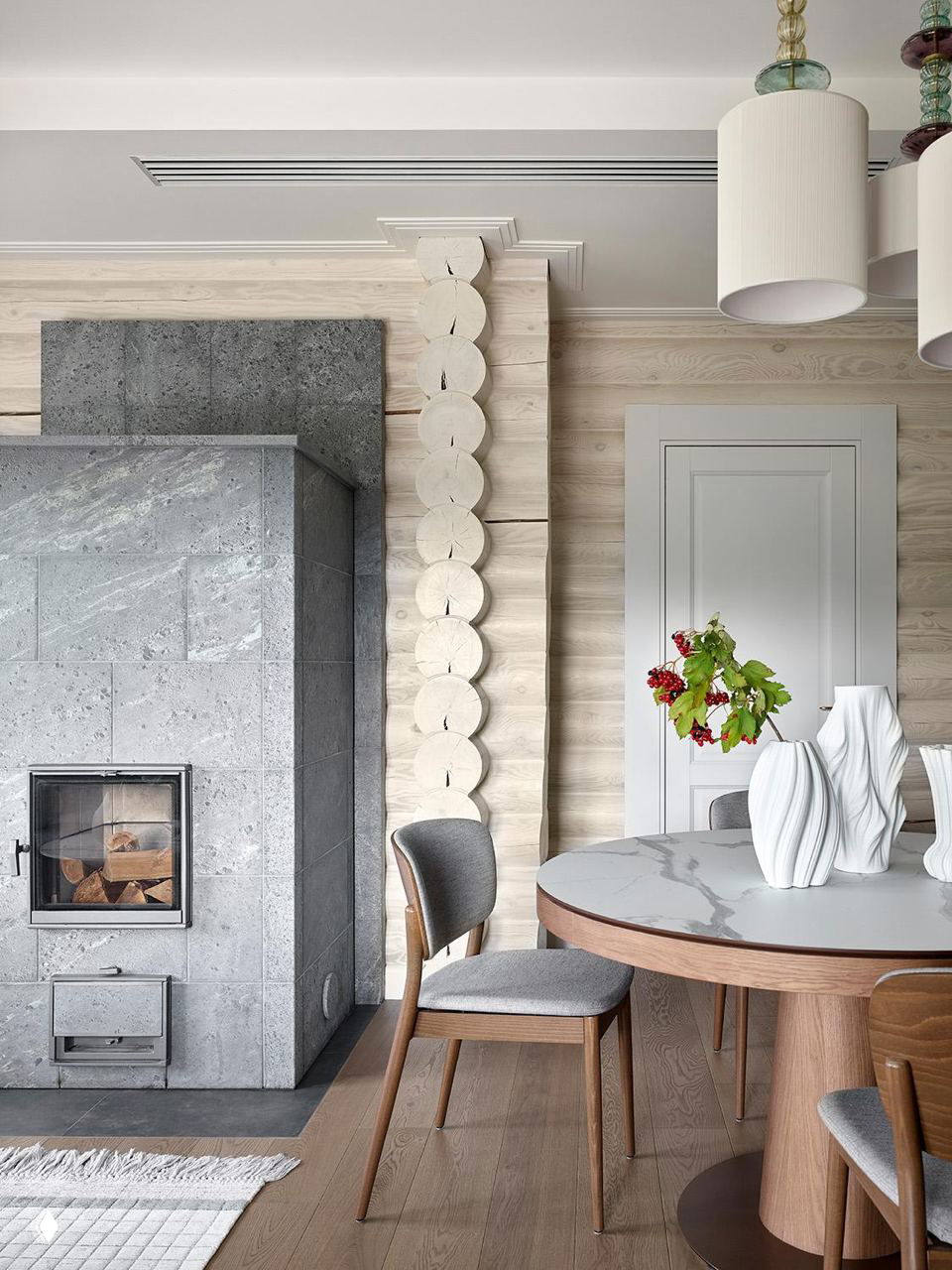Угол кухни-гостиной с камином из света серого камня, деревянной отделкой бревенчатых стен и круглым столом в зоне завтрака.