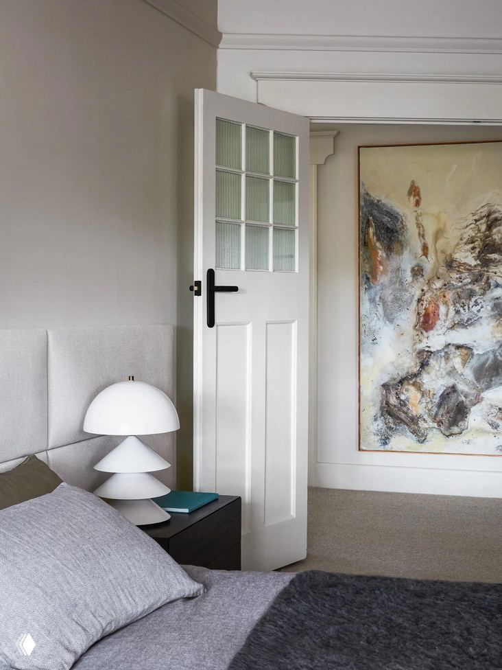 Спальня в викторианском доме: спокойная светлая палитра, низкая кровать с мягкой мебельной тканью и современный настольный светильник на тумбочке на фоне классической двери и большого абстрактного холста.