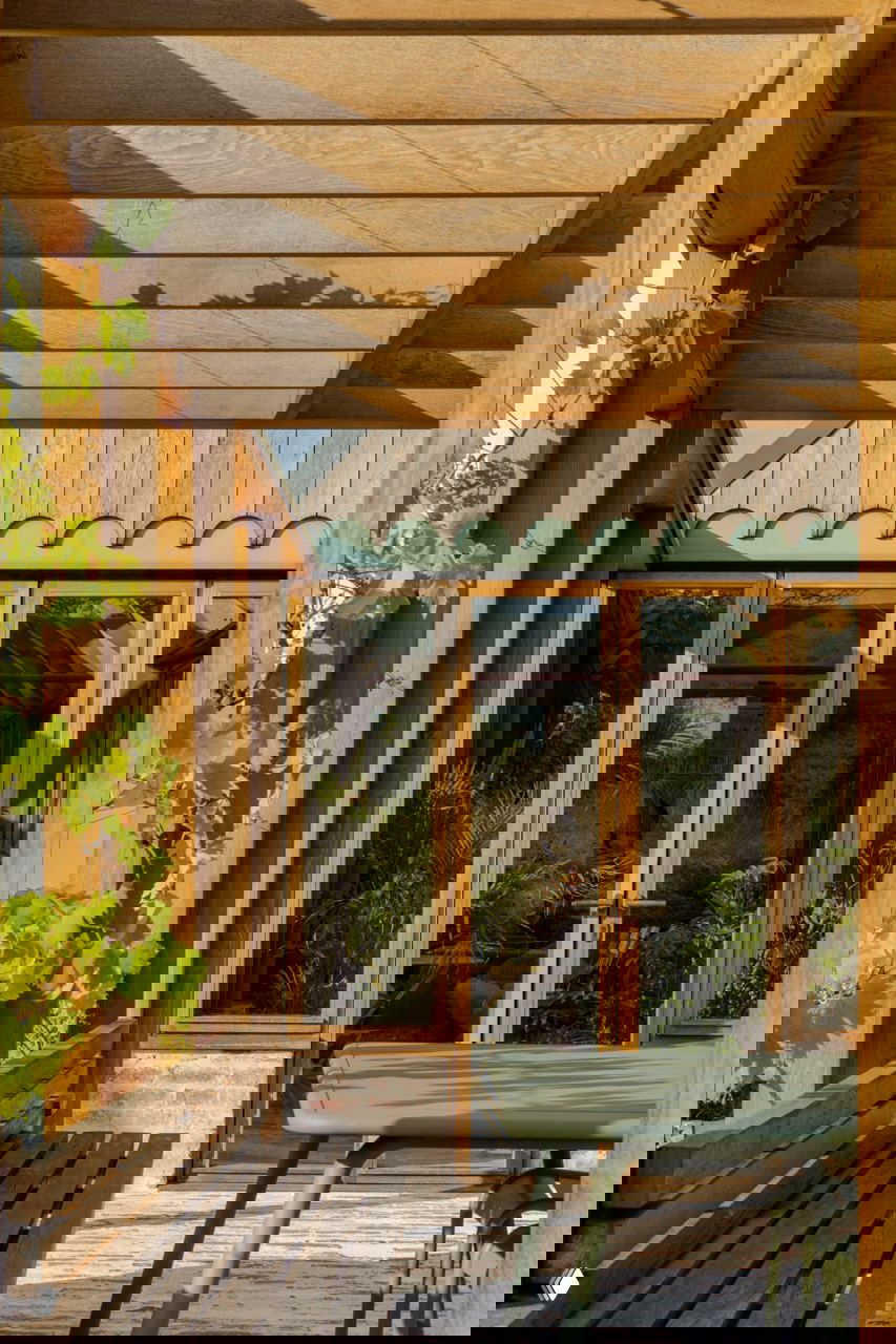 Улица и веранда с деревянными опорами, столовой зоной и большими стеклянными дверями, ведущими к зимнему саду с растениями.