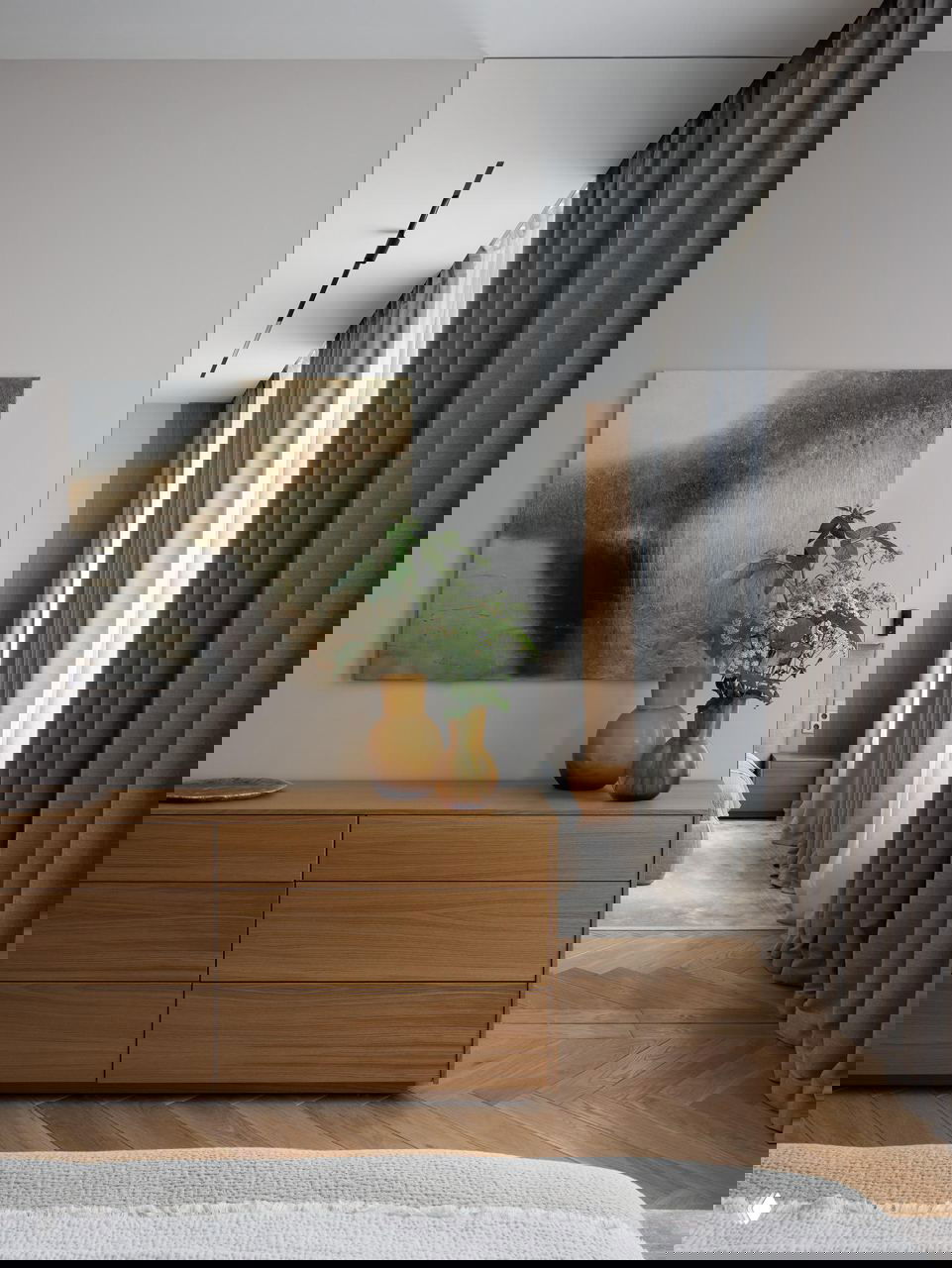 Деревянный комод с вазами и картиной, сквозной вид в спальню, длинные шторы и деревянный паркет в лаконичном интерьере.