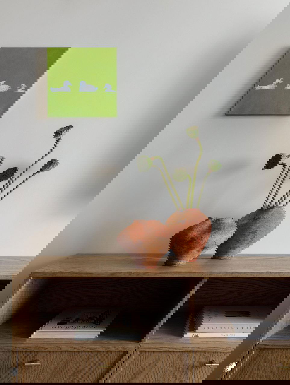 Композиция на тумбе: две керамические вазы терракотового цвета и зелёные стебли на фоне нейтральной стены и маленькой картины.