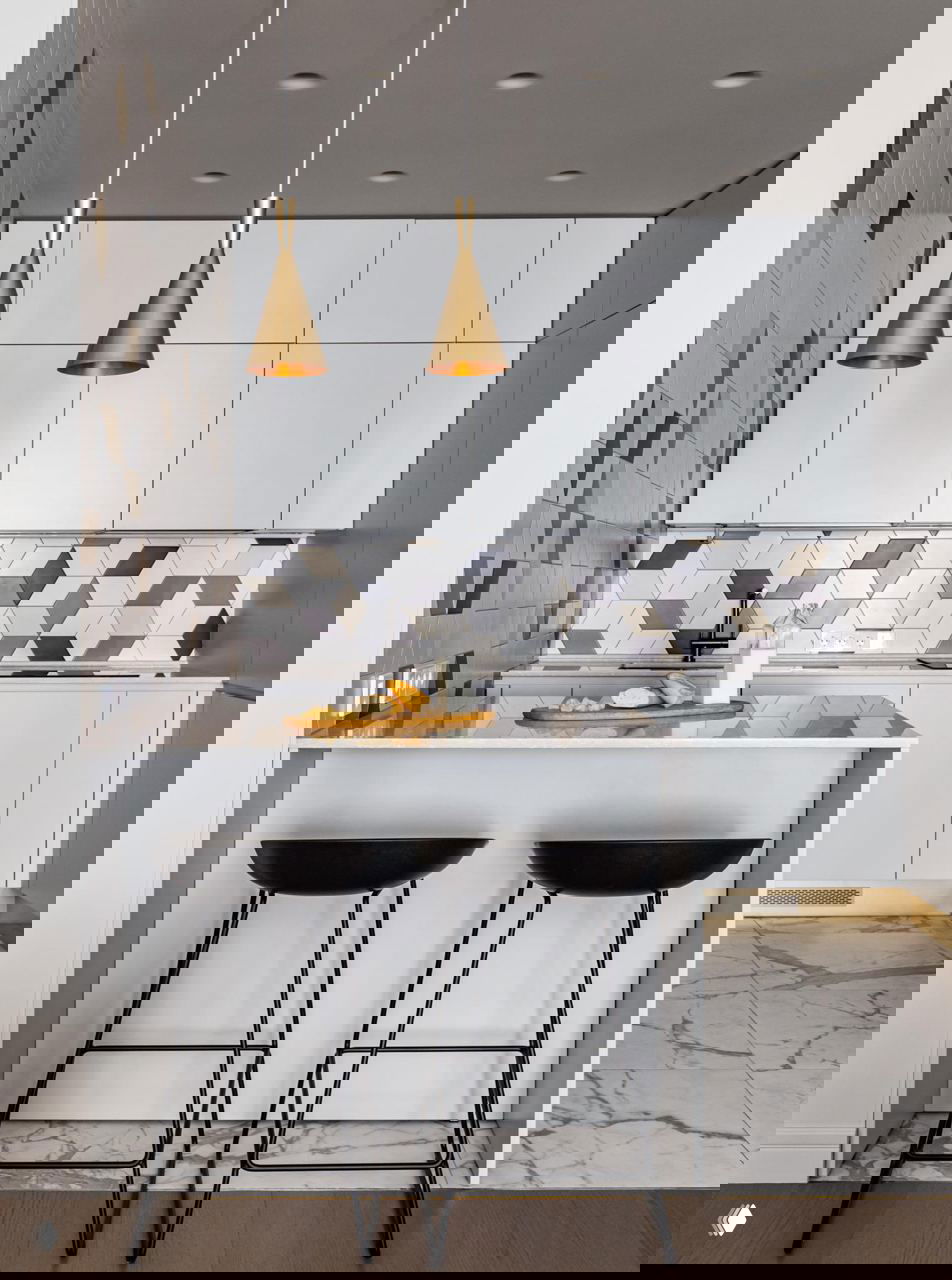 Светлая современная кухня с полуостровом и мраморной столешницей, двумя чёрными барными стульями и золотыми подвесными светильниками над рабочей зоной.