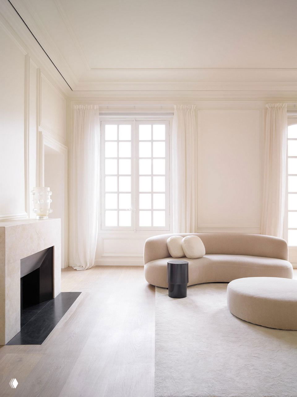 Просторная гостиная с бежевыми тонами, большим окном и минималистичной каминной нишей; мягкие текстуры и светлый пол подчёркивают монохромность.