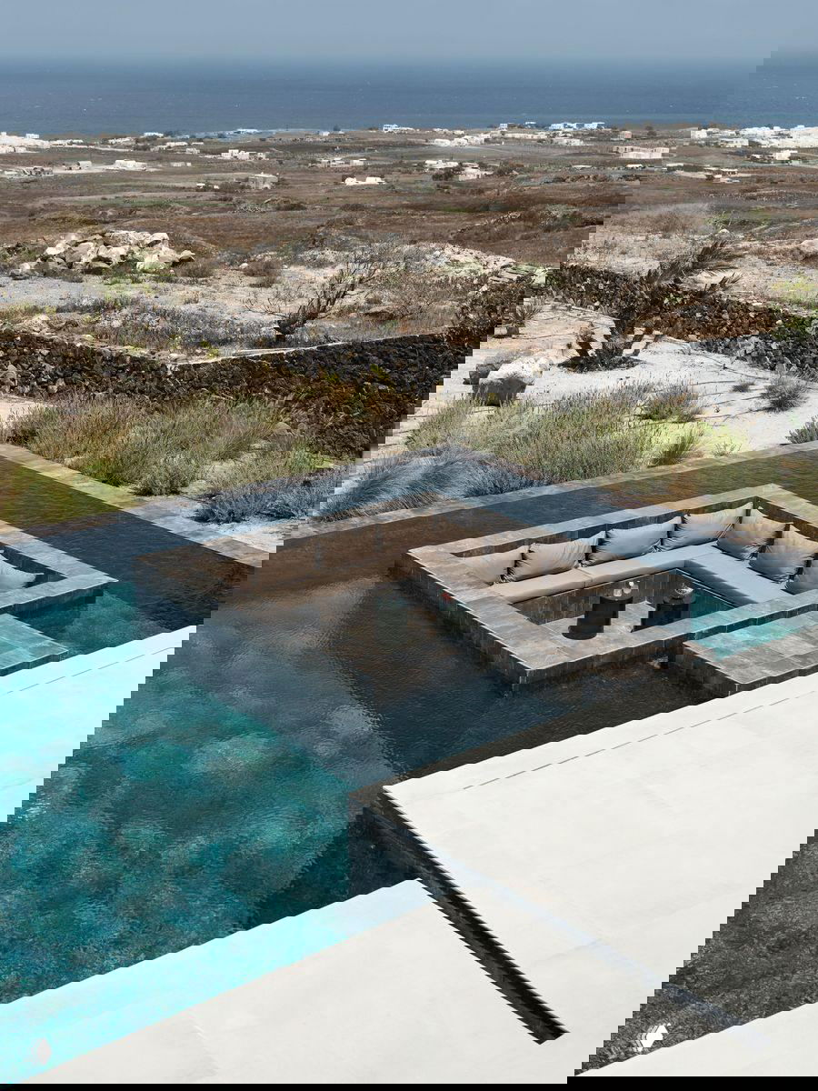 Встроенная в ландшафт открытая лаунж-зона в бассейне: угловой утопленный диван окружён водой, вид на море и каменистую местность на фоне.