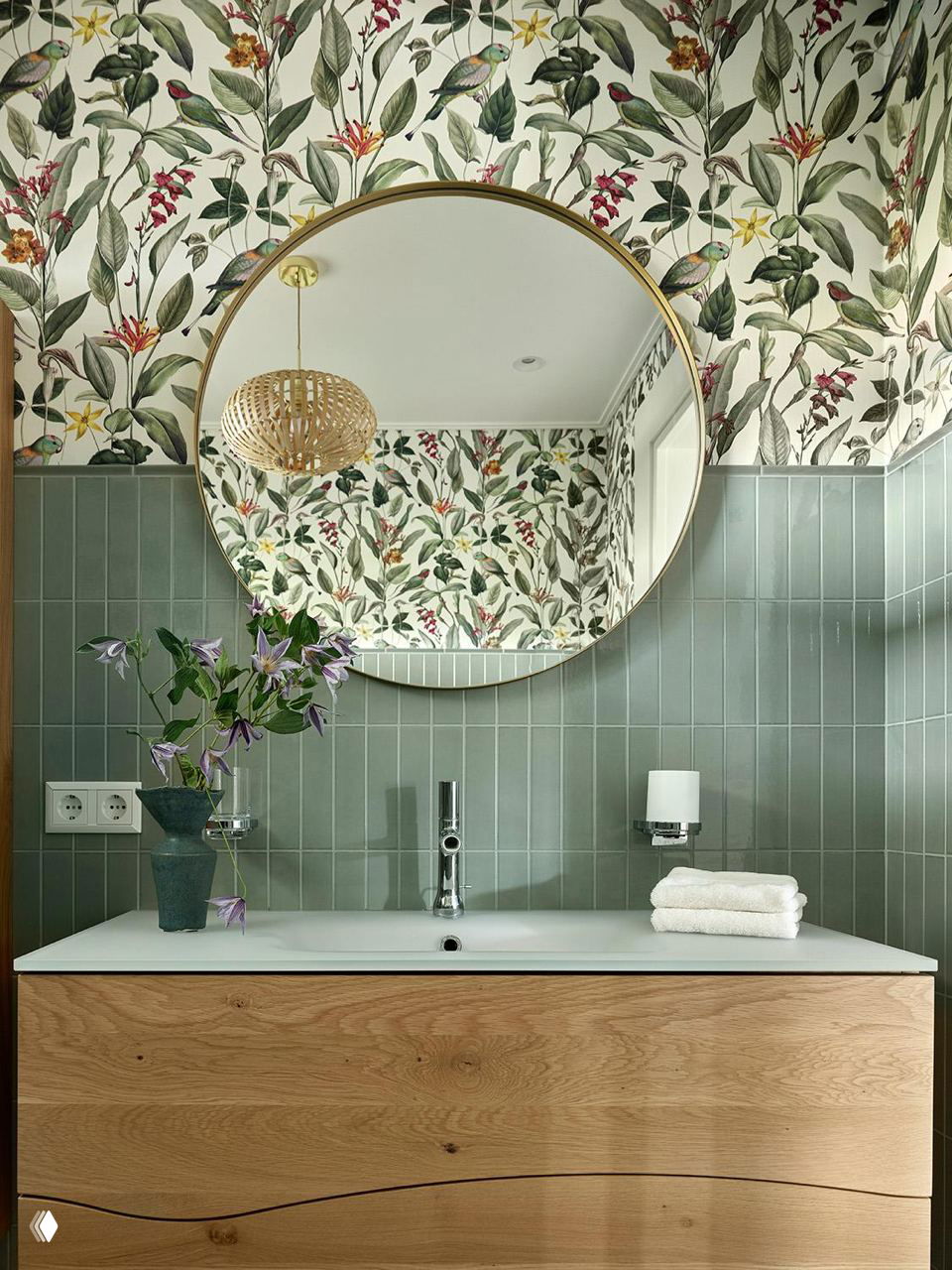 Ванная комната со стеклянным круглым зеркалом, зелёной плиткой и деревянной тумбой под умывальник на фоне флоральных обоев.