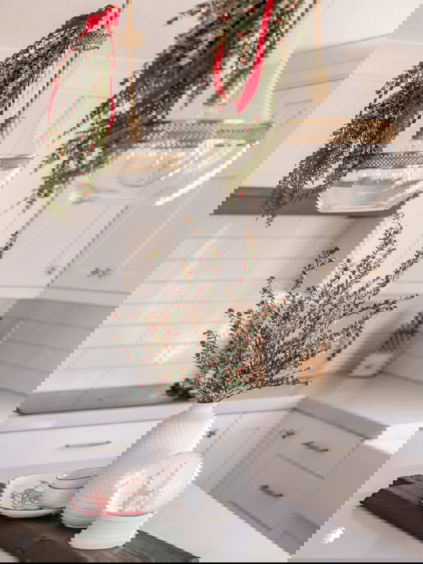 Подвесные стеклянные светильники, украшенные еловыми гирляндами и красными лентами над кухонным островом с декоративными ветками и подсветкой.