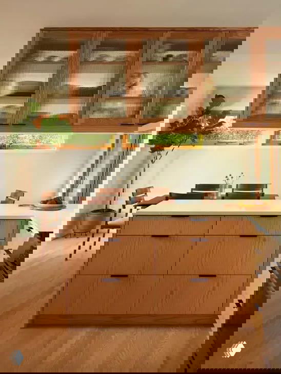 Тёплый интерьер с деревянным кухонным островом, выдвижными ящиками и подвесными шкафами со стеклом, создающий дополнительные места для хранения и визуальный объём.