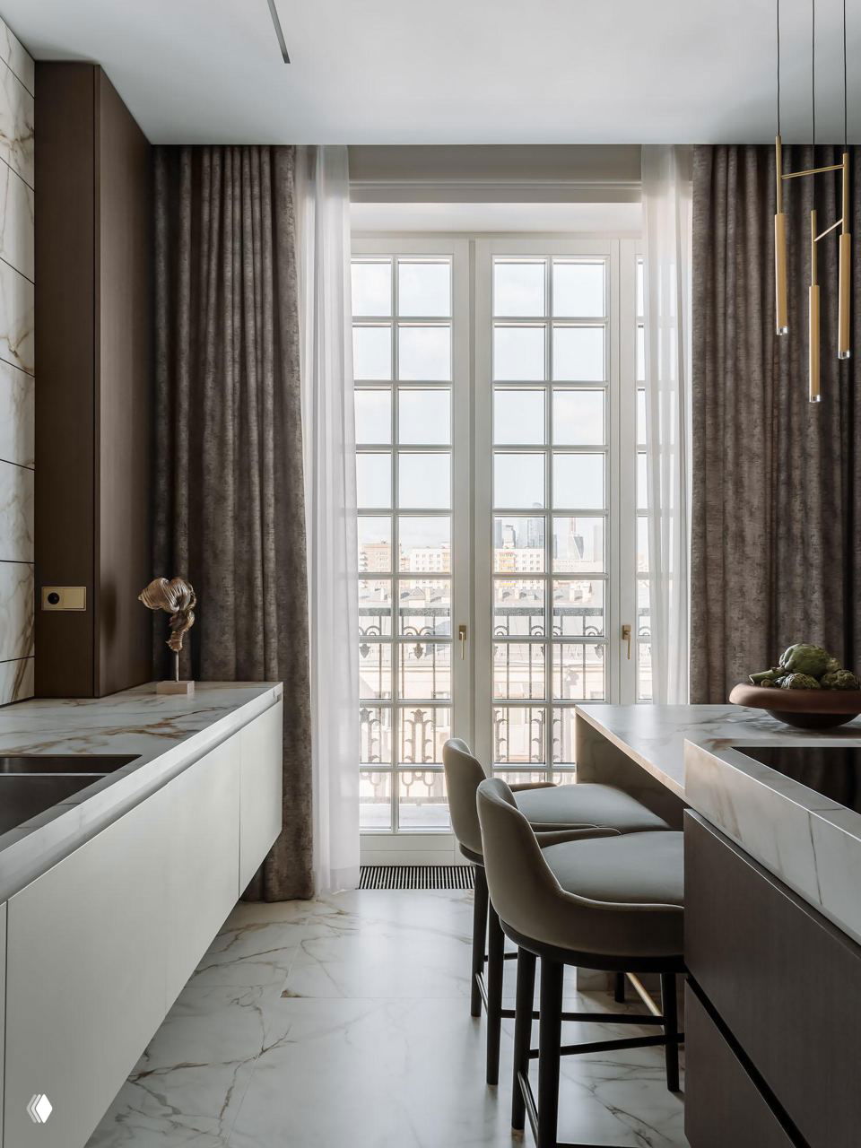 Кухня у французского окна: барные стулья у острова, мраморный пол, тёплые текстуры фасадов и прозрачные занавеси, пропускающие дневной свет в интерьер.