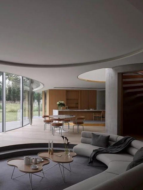 Круглая современная гостиная с низкой утопленной секцией и журнальными столиками, мягкие формы и большие панорамные окна создают свободное пространство.