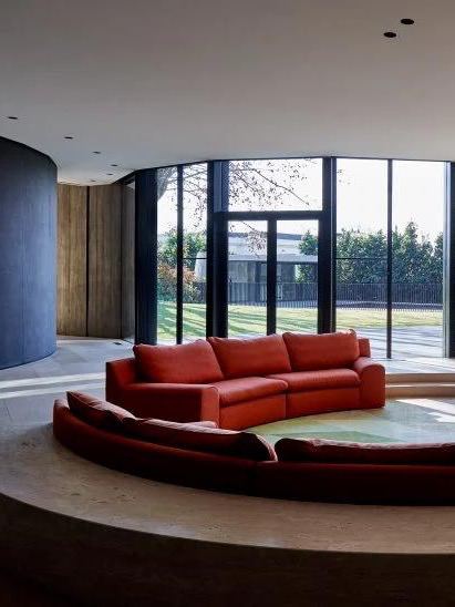 Утопленная в пол зона с красным модульным диваном на фоне панорамных окон и зелёного двора, сочетание округлых форм и больших стеклянных поверхностей.
