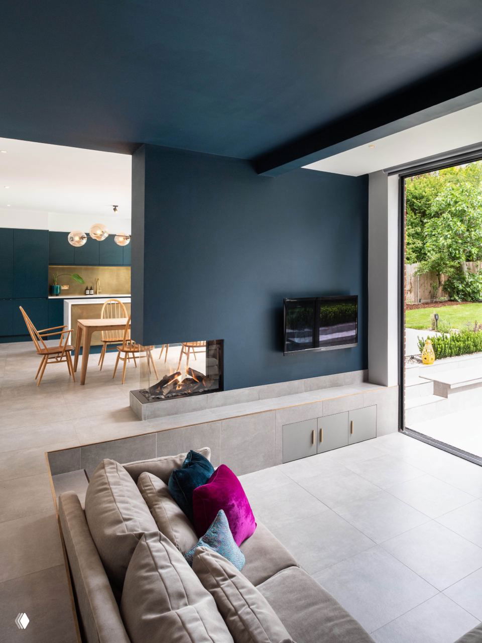 Современная утопленная гостиная с тёмно-синей акцентной стеной и камином в пониженном уровне, пространство объединено с кухней-столовой.